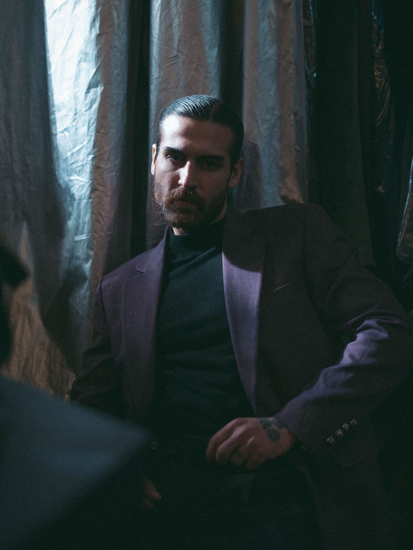 Man in a dark suit seated before heavy draped curtains