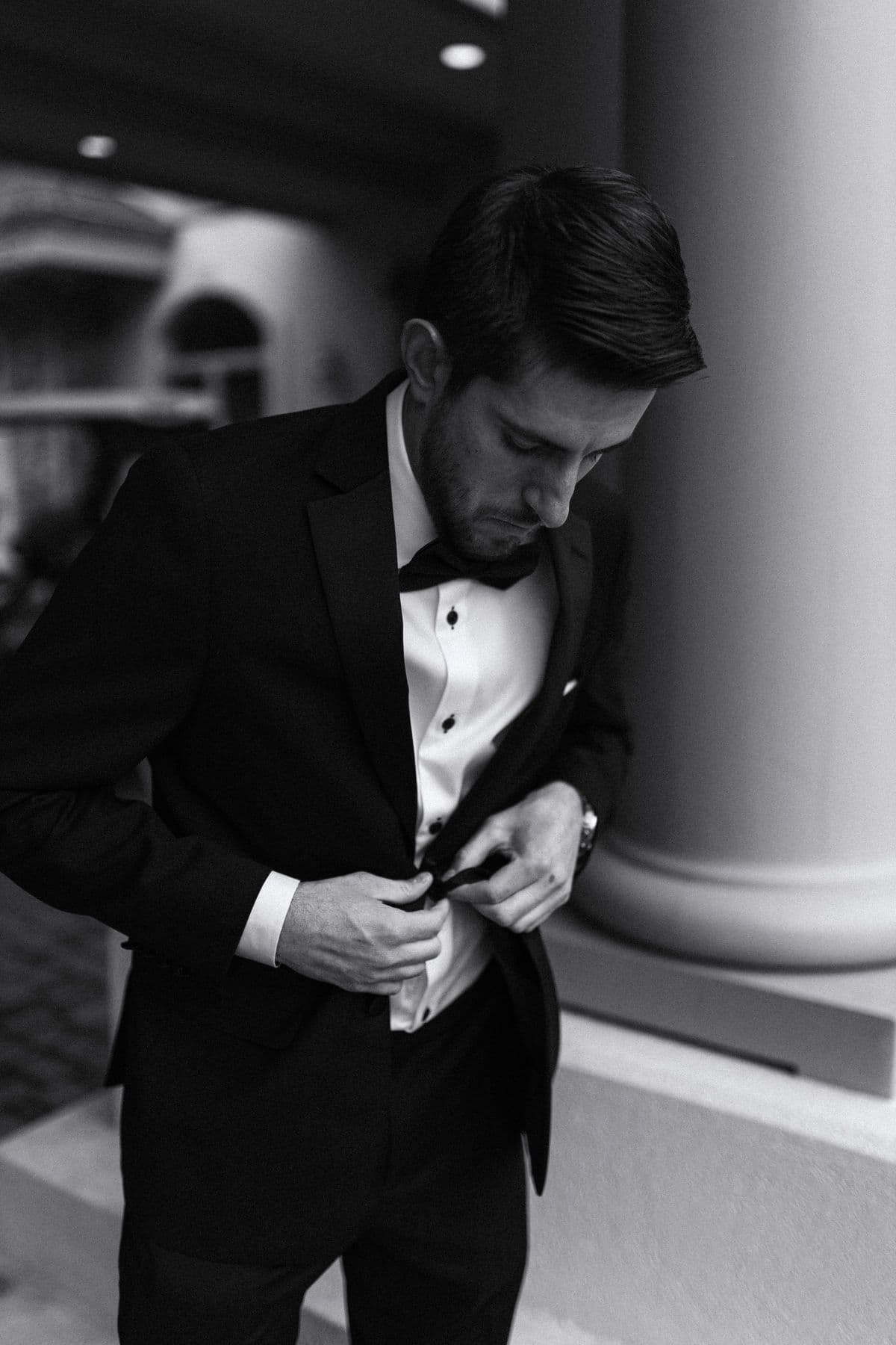 Black-and-white photograph of a groom in a tuxedo fastening his jacket beside a column