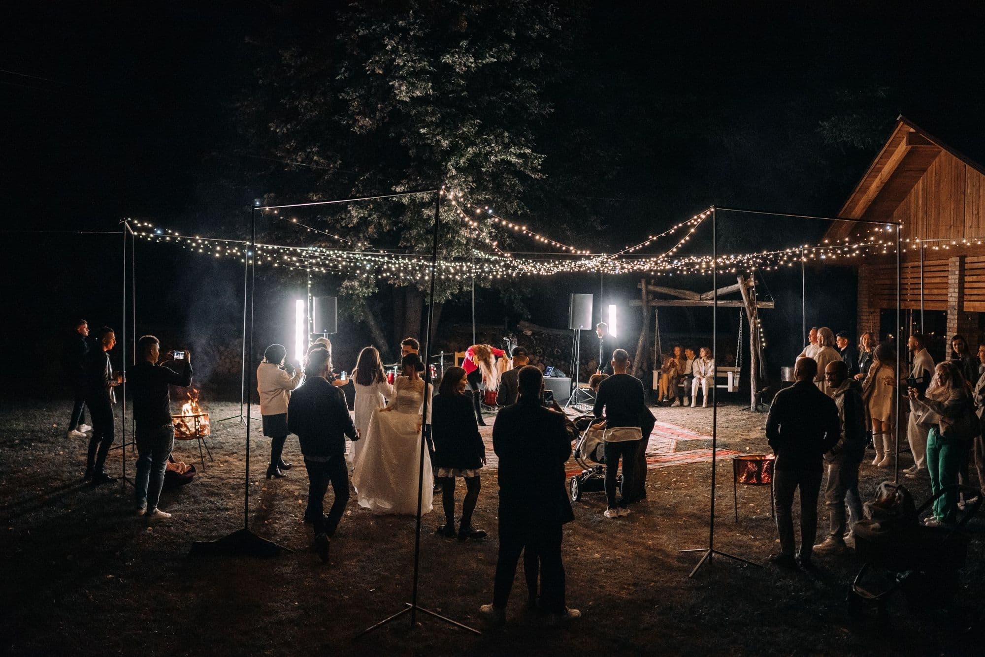 Guests gathered around the newlyweds at a nighttime outdoor reception beneath string lights beside a barn