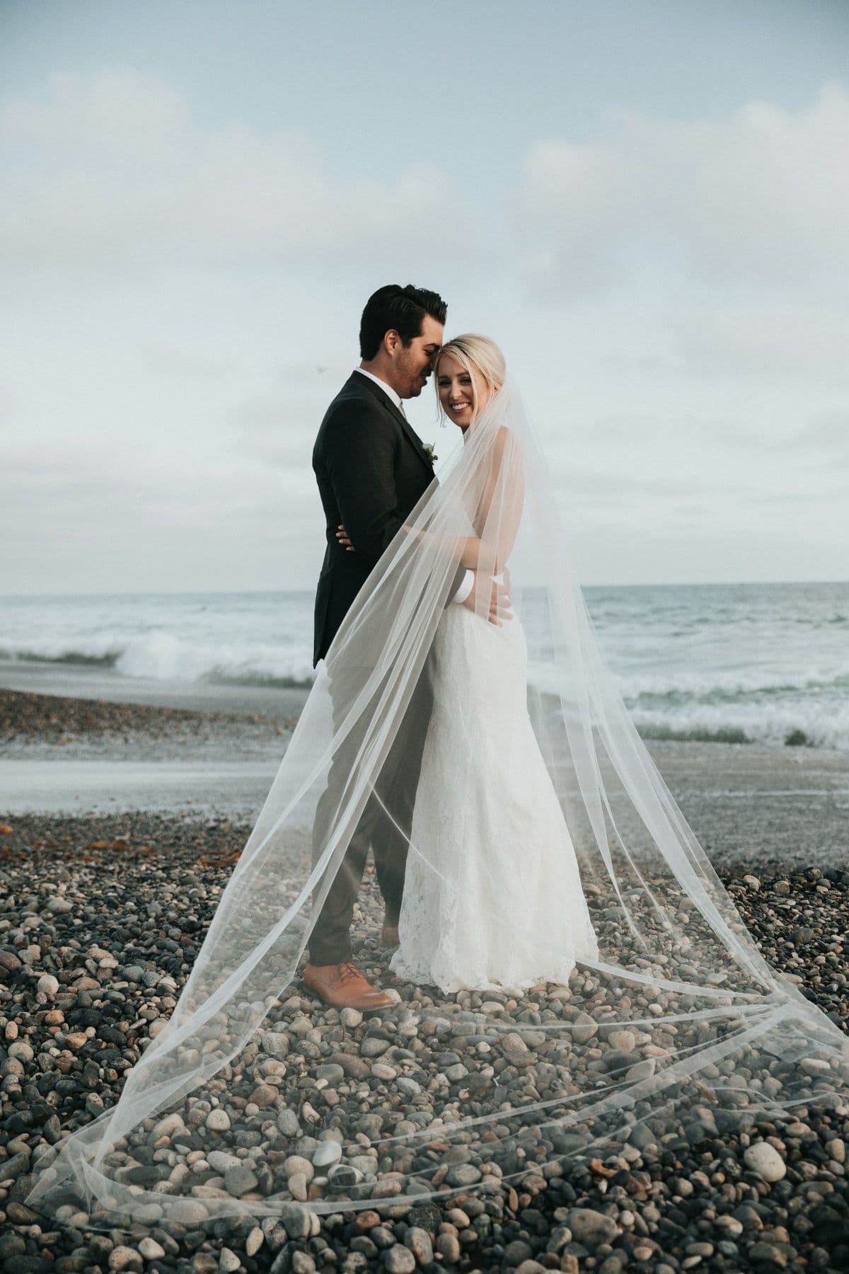 Bride and groom standing on a pebble beach as her cathedral veil drifts across the stones