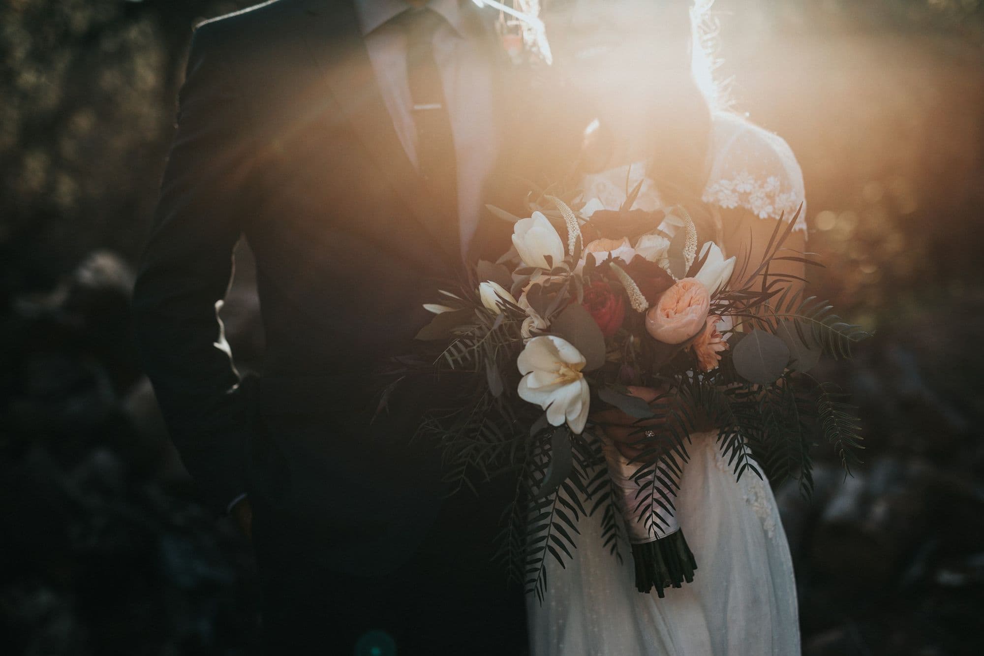 Groom holding his bride close as late sun flares across her bouquet of white and blush blooms