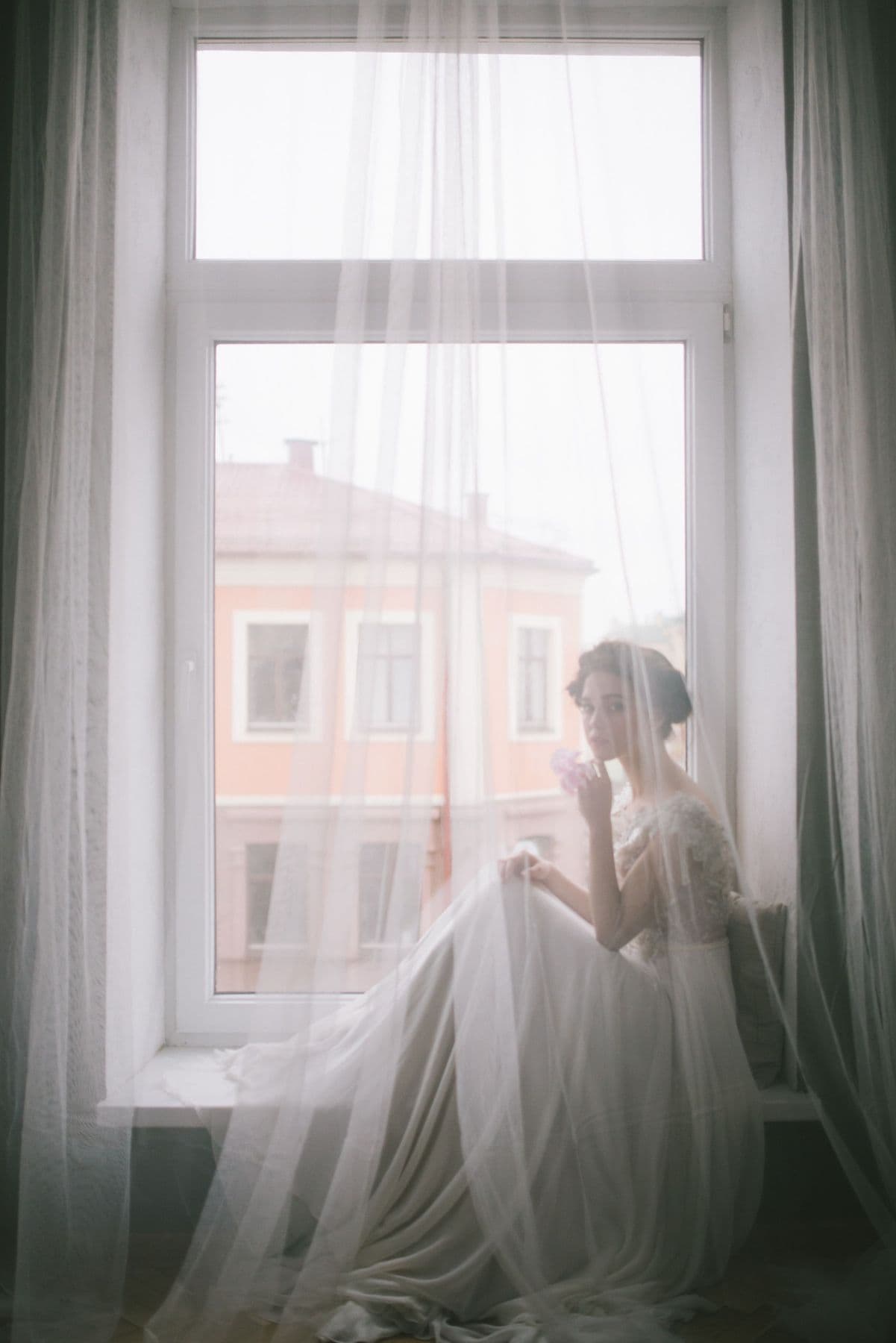 Bride in her gown seated beside a tall window with sheer curtains in soft morning light