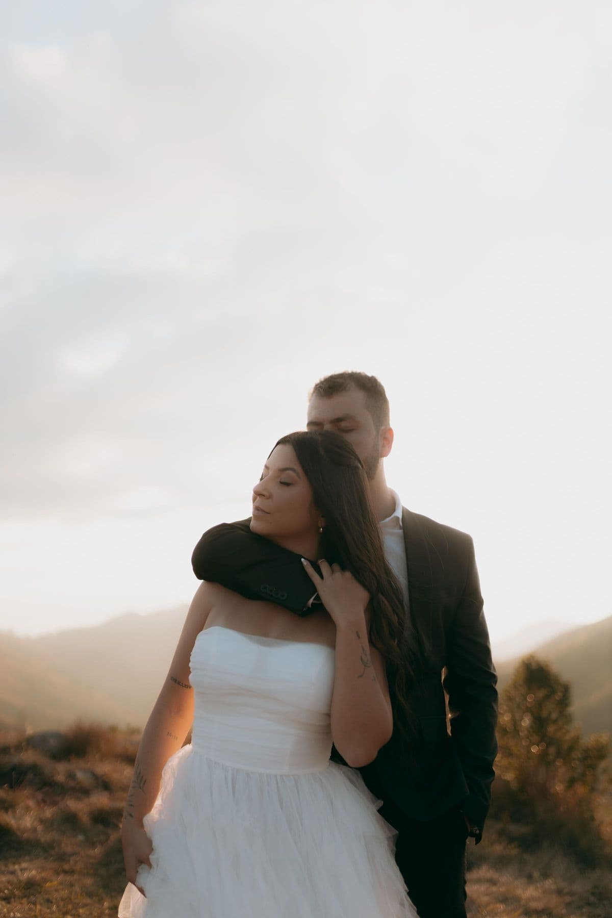 Groom embracing his bride from behind on a mountainside as the sun sets behind the ridge